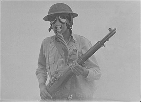 A soldier trains for fighting during a gas attack in WWI-era, flat-brimmed doughboy helmet and bolt-action rifle.